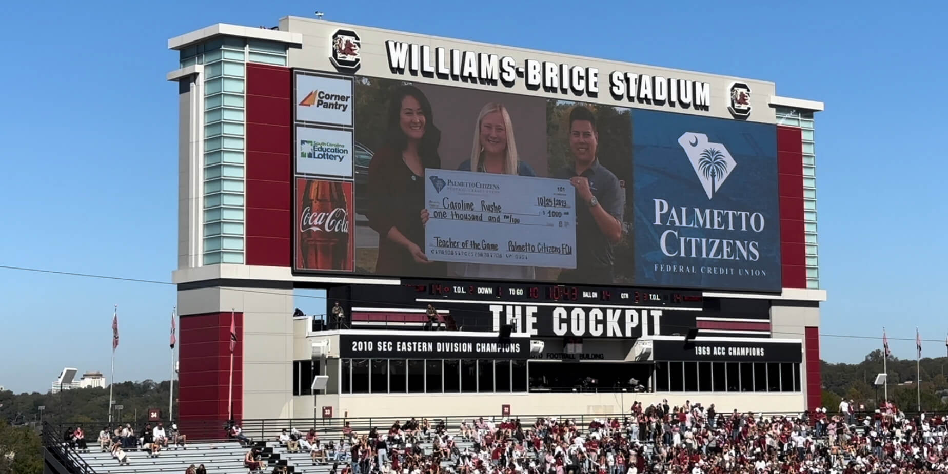 Scoreboard at Williams-Brice Stadium featuring Palmetto Citizens FCU's 'Teacher of the Game' with winners holding a large check.