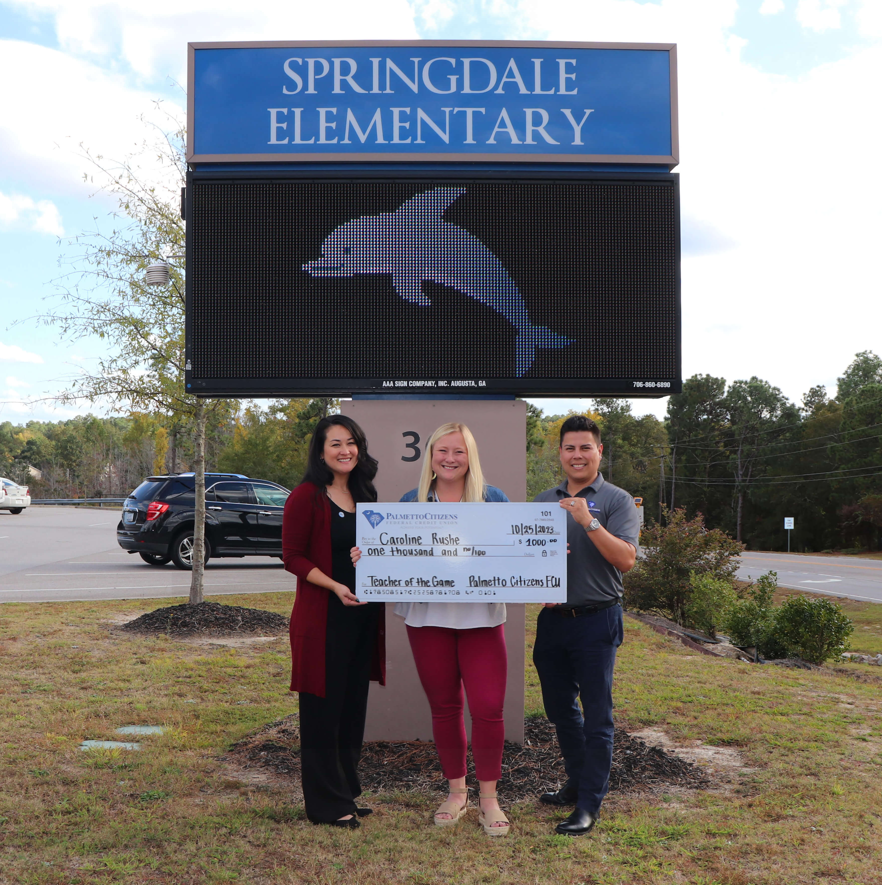 Three people posing with a 'Teacher of the Game' award check in front of Springdale Elementary's sign with a dolphin display.