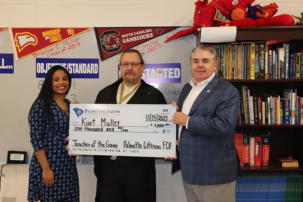 Three individuals in a classroom, with a man holding a large $1,000 'Teacher of the Game' check.