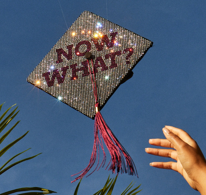 A hand throws a graduation cap in the air. The cap is bedazzled with the words Now what?
